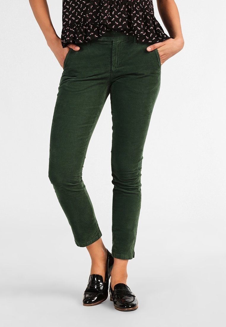 Femme portant un pantalon slim vert foncé, un chemisier noir à fleurs et des mocassins noirs brillants, les mains dans les poches.