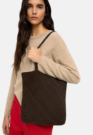 Kobieta o długich ciemnych włosach, ubrana w beżowy sweter i czerwone spodnie, trzyma na ramieniu dużą, ciemnobrązową plecioną torbę na ramię.