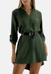 Groene jurk met V-hals, lange mouwen, omslagen aan de manchetten en een zwarte riem. Heeft een ontspannen pasvorm en een iets langere achterkant.