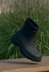 Bottine noire en caoutchouc avec une surface texturée, un haut incurvé et une semelle épaisse à crampons, présentée en lévitation au-dessus d'une surface en bois.