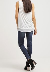 Top bianco senza maniche con orlo a strati, abbinato a jeans slim-fit blu scuro e stivaletti neri con tacco metallico.