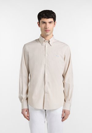 Giovane uomo che indossa una camicia beige a maniche lunghe con bottoni e pantaloni bianchi, in piedi davanti a uno sfondo bianco uniforme.