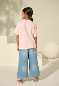 Helles rosa T-Shirt mit kurzen Ärmeln, kombiniert mit lässigen, weiten hellblauen Jeanshosen. Das Outfit hat eine lockere Passform und wird durch beigefarbene Sandalen ergänzt.