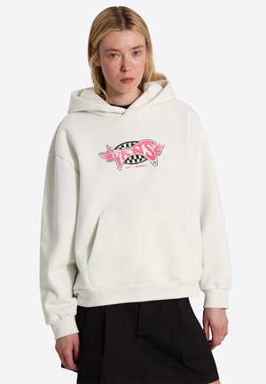 Ung person med lyst hår iført en hvid Vans-hættetrøje med rosa logo og sort nederdel, stående mod en ensfarvet baggrund.