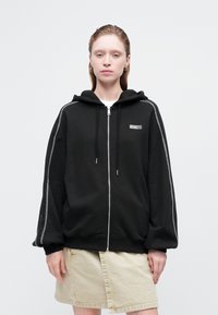 Mlada žena u crnoj zip-up dukserici s kontrastnim šavovima i bež suknji, stoji uz običnu svijetlu pozadinu.