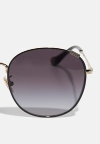 Óculos de sol com uma forma grande e arredondada, lentes escuras em degradé, armação metálica fina em dourado e hastes pretas. Apresenta um logótipo na armação.