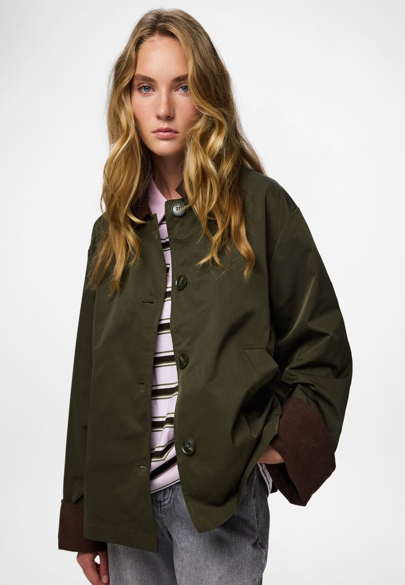 Oliivroheline bomberjakker, millel on ruudukujuline vorm, suured nupud ja pruunist korduroost mansetid. Kantud triibulise särgi ja hallide teksade üle.