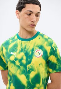 Νεαρός άνδρας με κοντά σκούρα μαλλιά που φοράει φανέλα ποδοσφαίρου της Σενεγάλης σε πράσινο και κίτρινο tie-dye, με λογότυπα Puma και της ομάδας.