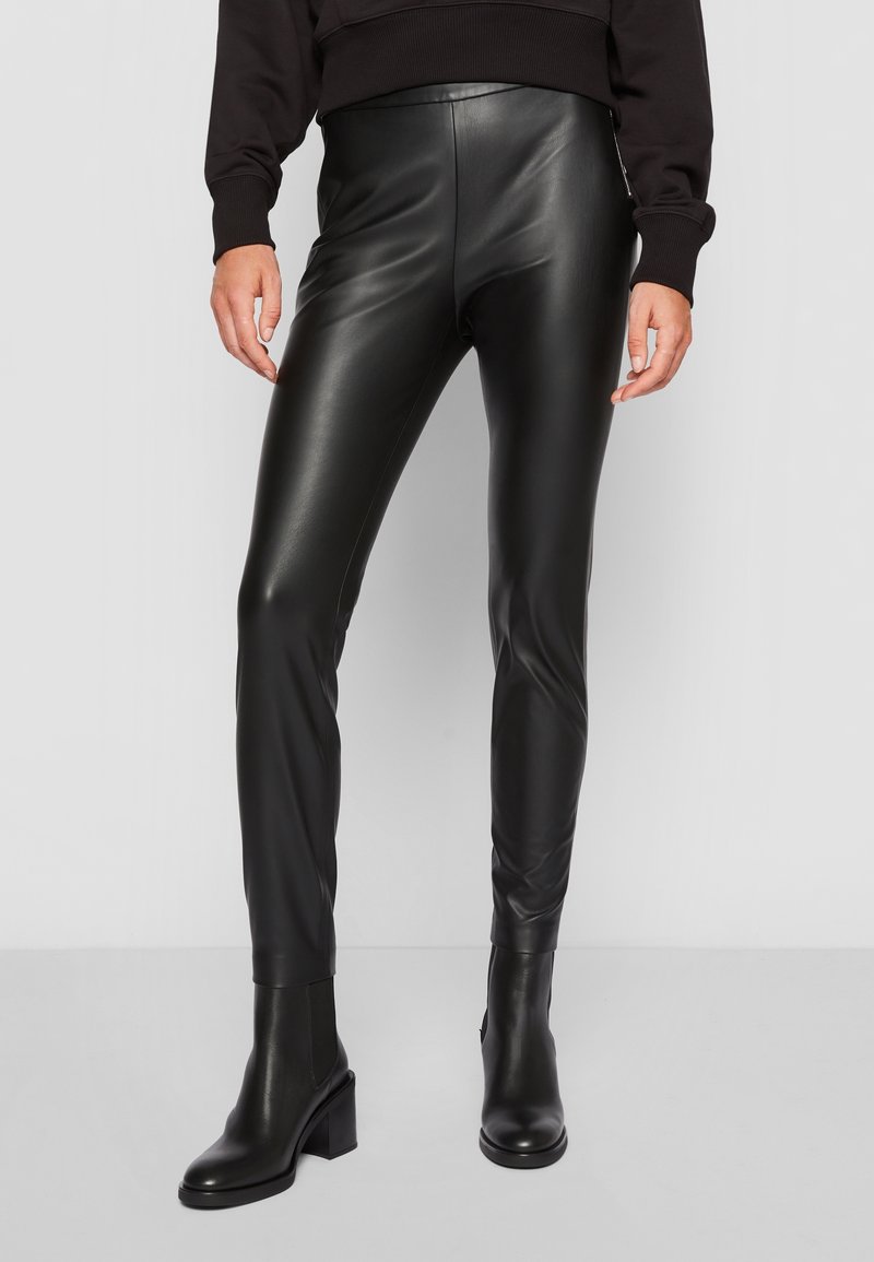 Personne portant un pantalon noir moulant en similicuir et des bottines à talons noires, posant devant un fond clair uni.