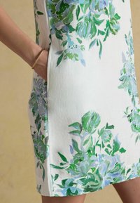 Hand in pocket of white dress with green and blue floral pattern, person wearing gold bracelet, beige background.