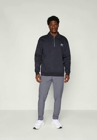 Felpa navy a zip con colletto a coste e polsini a coste, abbinata a pantaloni leggeri grigi a gamba affusolata. Le scarpe sono un paio di sneakers bianche.