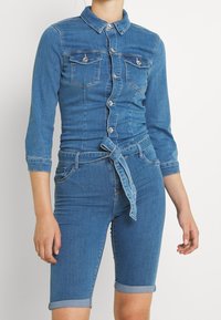 Kvinna iklädd en figursydd blå denimskjorta med knäppning och bröstfickor, kopplad med ett knytskärp, i kombination med knälånga uppvikta denimshorts.