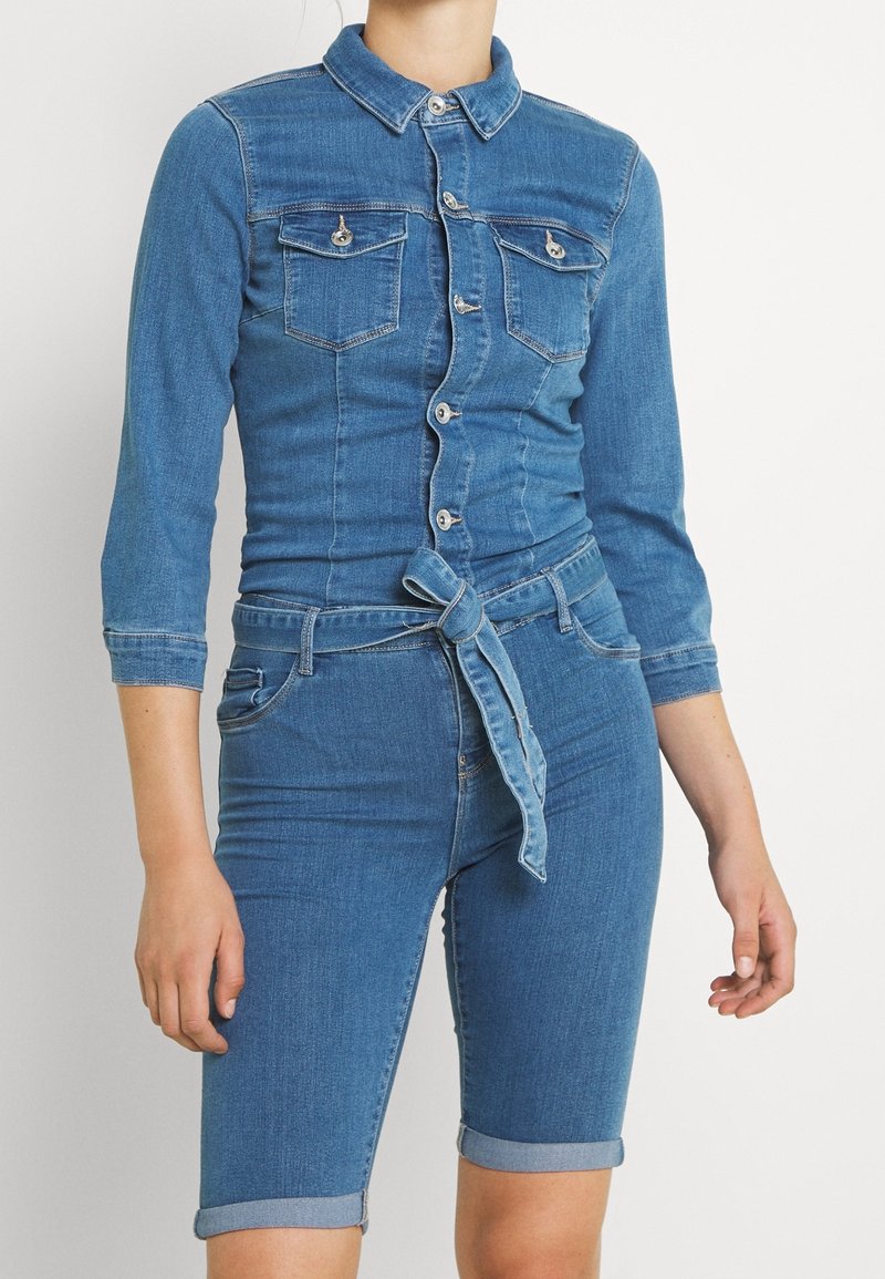 Kvinna iklädd en figursydd blå denimskjorta med knäppning och bröstfickor, kopplad med ett knytskärp, i kombination med knälånga uppvikta denimshorts.