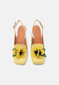 Κίτρινα παπούτσια με λουράκι και πουά, με τετράγωνη μύτη, διακοσμημένα με τσόχινα λουλούδια σε κίτρινο και μαύρο, και καφέ υφασμάτινη εσωτερική σόλα.