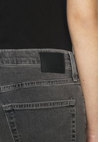 Jean en denim gris avec une surface texturée, comportant une poche en cuir noir à l'arrière et des coutures le long des poches.