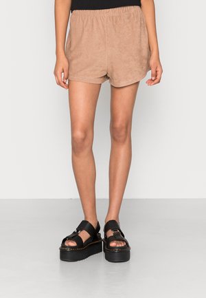 Person iført beige ruskindsshorts og sorte platformsandaler, stående mod en ensfarvet lys baggrund.
