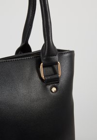 Sac à main en cuir noir avec une texture lisse, doté d'accents métalliques dorés, d'un design structuré et de deux poignées robustes.