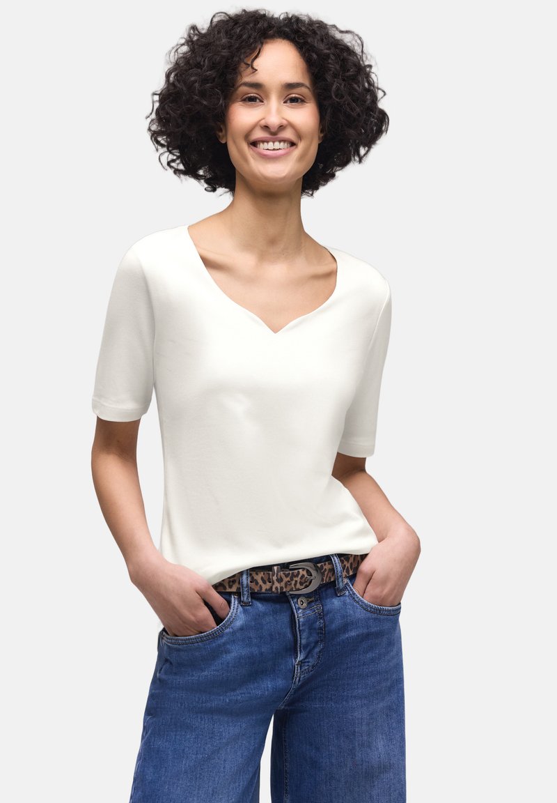 Femme aux cheveux bouclés souriante portant un haut blanc à manches courtes, un jean bleu et une ceinture à imprimé léopard, les mains dans les poches, sur un fond uni.