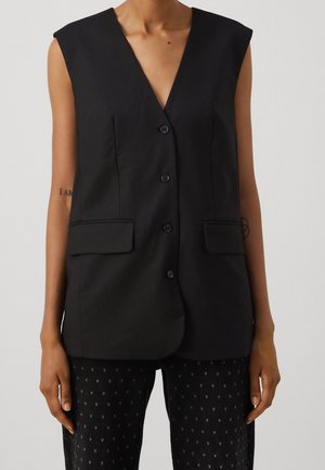 Persoon draagt een mouwloos zwart knoopvest met twee voorflappzakken en een zwarte broek met patroon, staand tegen een effen achtergrond.