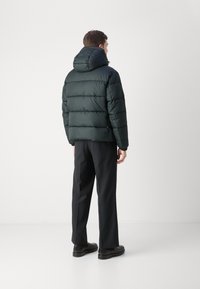 Dunjakke i mørkegrønn med hette, med et quiltet design og elastiske mansjetter. Brukt med svarte rettbukser og mørke sko.