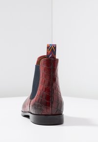 Bottine en cuir à motif crocodile rouge avec un panneau latéral élastique noir et une languette en tissu multicolore à l'arrière sur une surface blanche.