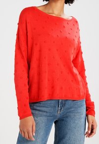 Roter gestrickter Pullover mit lockerem Schnitt, rundem Ausschnitt und strukturiertem Punkte-Muster. Kombiniert mit blauen Jeans.