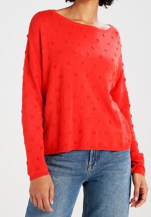 Rød strikket sweater med løs pasform, rund halsudskæring og tekstureret prikmønster. Sammen med blå denimjeans.