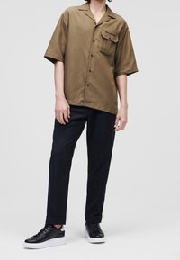 Olijfgroene shirt met korte mouwen, een voorkantzak, knoopdetails en een losse fit, gecombineerd met zwarte broeken en zwarte sneakers met witte zolen.