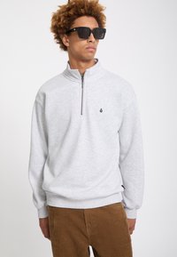 Jeune homme aux cheveux bouclés portant des lunettes de soleil noires, un sweat-shirt gris clair à fermeture éclair sur le quart et un pantalon en velours côtelé marron, debout devant un fond blanc.