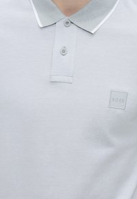 Polo gris clair avec deux boutons, un col côtelé avec bordure blanche, et un petit logo carré embossé « "BOSS" » sur la poitrine.