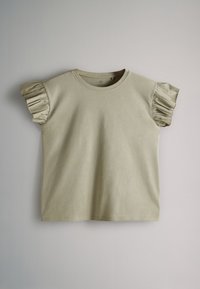 Lyse beige t-shirt med korte ærmer, rund hals og flæsedetaljer på ærmerne, lagt fladt på en neutral baggrund.
