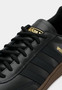 Μαύρο δερμάτινο sneaker με τρεις σκούρες μπλε ρίγες, χρυσό λογότυπο "Adidas", μαύρες κορδόνες και καφέ λαστιχένια σόλα.