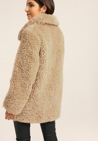 Cappotto in pelliccia sintetica beige con un ampio colletto, superficie texturizzata e vestibilità ampia, abbinato a pantaloni neri, mostrato da dietro.