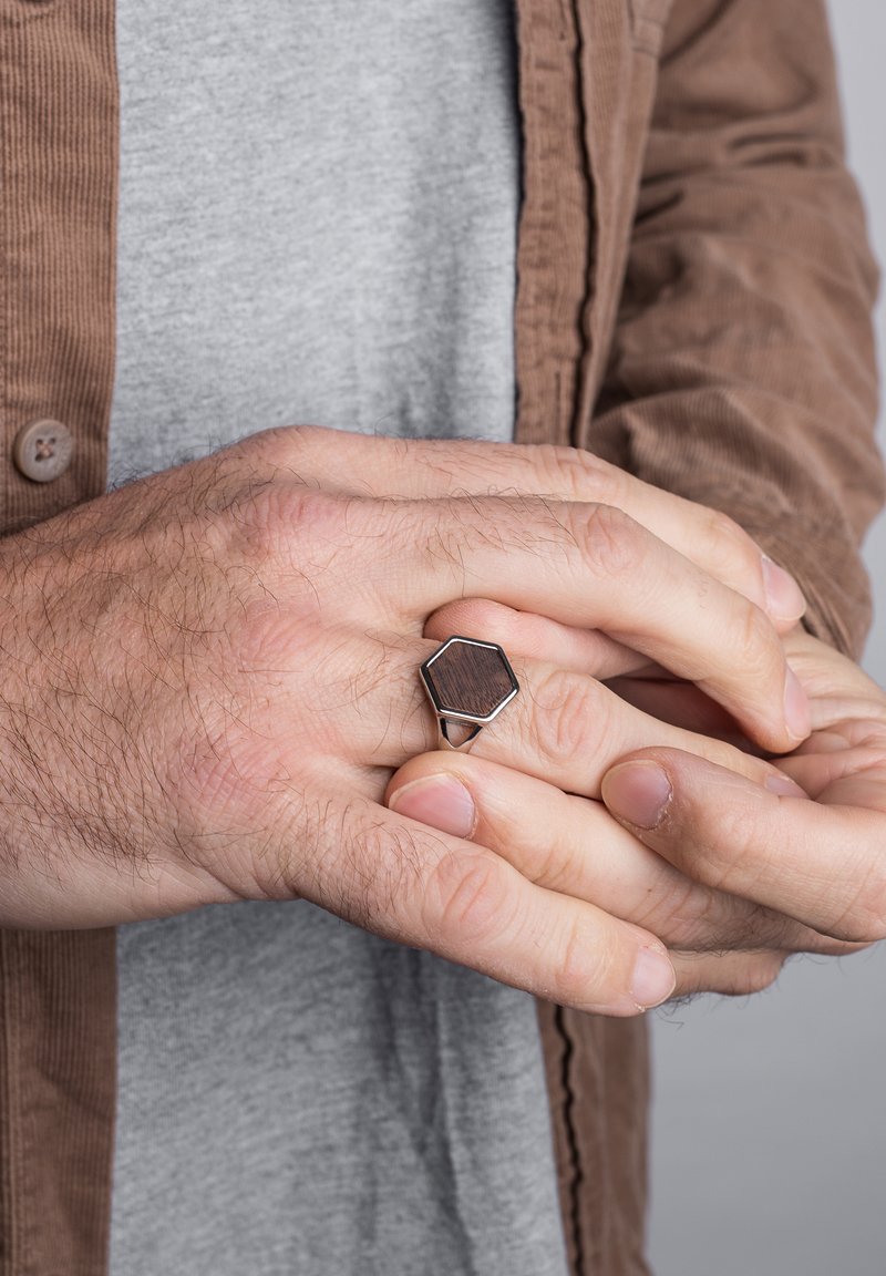 Zilveren hexagonale ring met een donkere houten inzet, gedragen op een hand met zichtbare haartjes, tegen een grijze achtergrond en neutrale kleding.