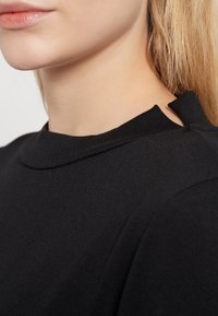 Svart topp med hög hals och gradvis axelutskärning, med en slät textur och minimalistisk design.