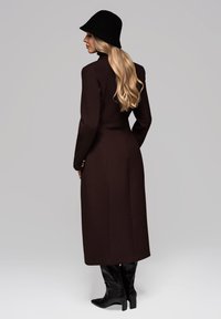 Brun uldblanding lang frakke med tætsiddende silhuet, høj krave og gyldne knapdetaljer. Sat sammen med sorte læderknæstøvler og en sort hat.