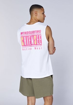 Mann in weißem ärmellosem Shirt mit pinkem und gelbem Aufdruck „Windsurfing Chiemsee Active Wear“, trägt olivgrüne Shorts und steht mit verschränkten Armen.