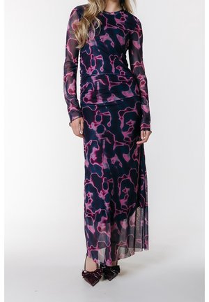 Femme portant une robe longue à manches longues jusqu'au sol avec des motifs abstraits bleu foncé et rose et des chaussures brillantes foncées à bouts pointus avec de grands nœuds.