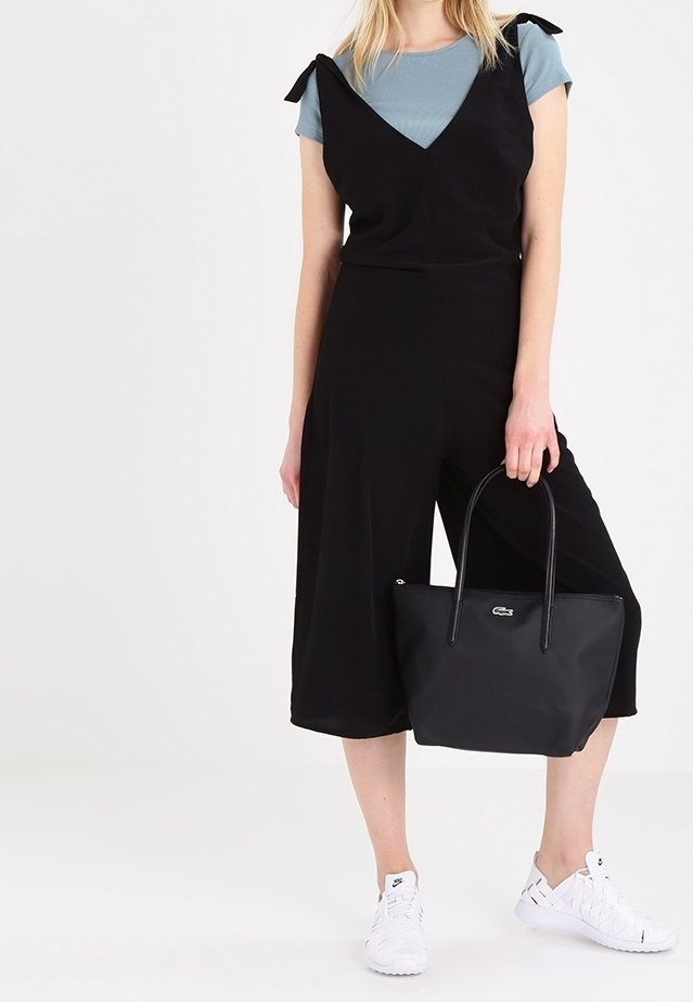 Femme portant une combinaison noire sans manches par-dessus un t-shirt bleu, tenant un sac fourre-tout noir et portant des baskets blanches, debout sur un fond blanc.