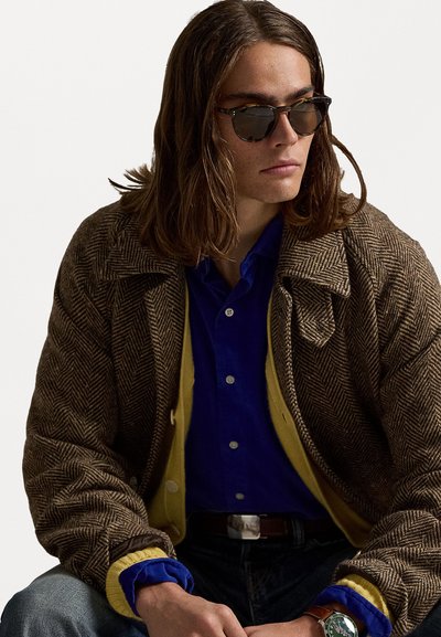 Ungdom med langt hår iført mørke solbriller, lagdelt brun herringbone-kåpe, blå skjorte, gul cardigan, jeans og et grønt armbåndsur.