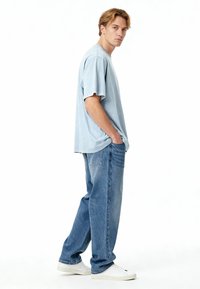 Ljusblå oversized t-shirt med korta ärmar, tillsammans med avslappnade denimjeans. Modellen bär vita sneakers. En enkel bakgrund.