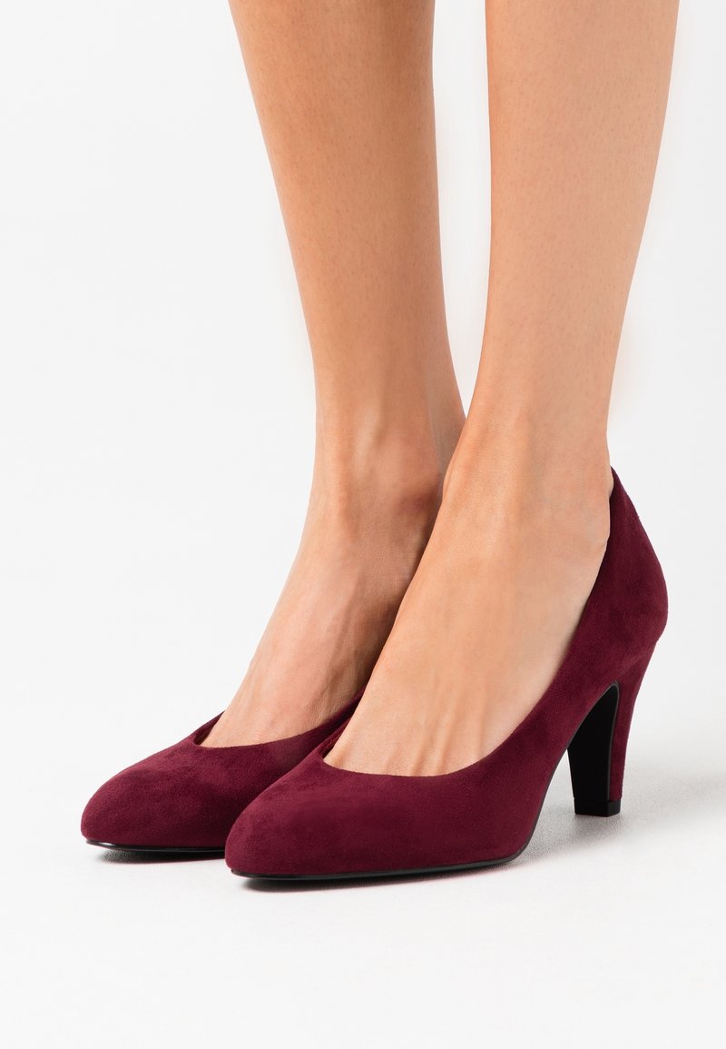 Paire de jambes portant des escarpins en daim bordeaux à bout fermé arrondi et talons fins sur fond blanc.