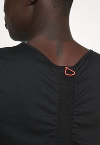 Närbild av en persons övre rygg iförd en svart topp med samlad mittsöm och en liten orange ögla nära halsringningen.