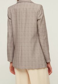 Blazer a quadri marrone chiaro con un design aderente, spalle strutturate e una texture liscia, dotato di un singolo spacco posteriore e maniche lunghe.