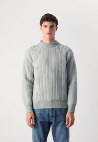 Light green ribbed knitted sweater with a high crew neckline and textured pattern. Soft fabric, relaxed fit, paired with blue denim jeans.