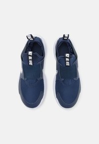 Baskets de sport bleu marine avec une tige en maille texturée, une sangle élastique et une semelle blanche. La languette au niveau du talon présente un logo.