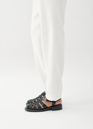 Άτομο που φοράει λευκό παντελόνι και μαύρα δερμάτινα σανδάλια ανοιχτής μύτης με αγκράφα, στέκεται σε λευκό φόντο.