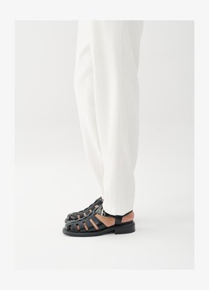 Isik, kes kannab valgeid pükse ja musta nahast avatud varvastega sandaale pandlaga, seisab valge tausta ees.