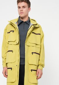 Casaco exterior amarelo claro com múltiplos bolsos frontais, fechos de correr pretos e capuz. Sobreposto a um pulôver cinza escuro.