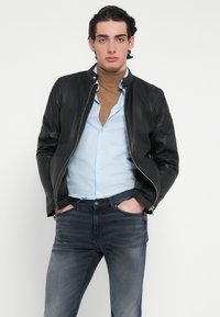 Jachetă bomber neagră din piele cu guler tip stand, închidere cu fermoar, purtată peste o cămașă albastru deschis și un pulover maro cu guler înalt, asortată cu blugi închiși la culoare.